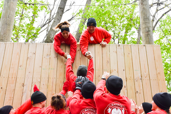 Two recruits are leaning over the top of a tall wall, and are pulling up a third recruit. A group of recruits at the bottom of the wall are helping left up the recruit.