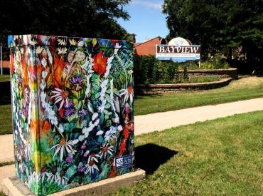A square utility box is covered in a multi-color watercolor painting of flowers and grasses. The box is in front of a sidewalk, adjacent to a community center with a sign that reads “Bayview Foundation”