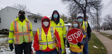 Image of four masked crossing guards in yellow vests holding a red stop sign.
