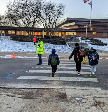 Imagen de 3 personas cruzando en un paso de peatones hacia un guardia de cruce con chaqueta amarilla que mantiene detenido el tráfico.