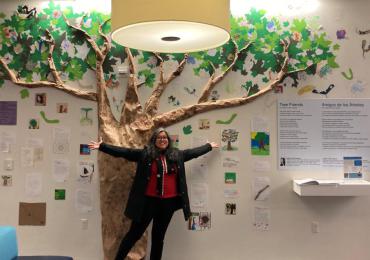 A woman with dark hair, in a red sweater with a black jacket stands with her arms outstretched in front of a wall depicting a tree and dozens of poems under the tree on different colored and sized pieces of paper. 