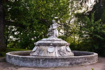 The Annie Stewart Fountain is shown in its current condition with black lichen on the once white marble sculpture. The Central figure is a mermaid who pours water  into a shell held by a child.