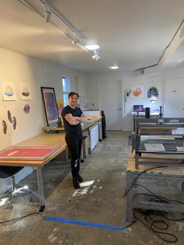 An individual in black pants and a black shirt stands with their arms crossed smiling in a room full of art supplies and art, including a print press and screen printing screens,