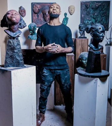 An artist in black clothes stands with his hands clasped in front of a a collection of figurative sculptures.
