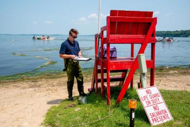 A person wearing waders standing on a beach next to a red lifeguard chair, holding a clipboard and pen