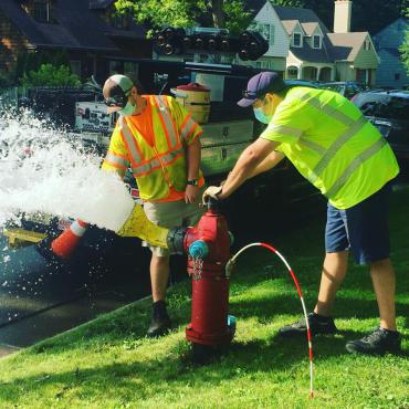 Two Madison Water Utility employees wearing masks and safety vests are working at a fire hydrant. Water sprays from the hydrant as one employee opens it and the other collects a water sample.