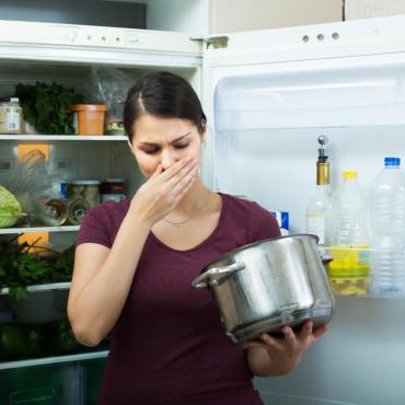 Woman standing in front of open refrigerator with cooking pot in hand, holding hand over nose