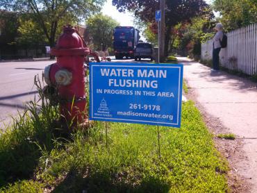 A blue sign in a terrace reads: "Water main flushing in progress in this area. 261-9178 madisonwater.org"