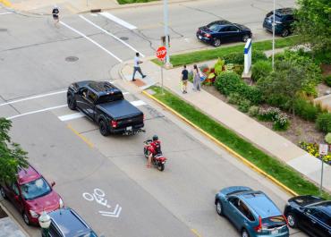 Imagen que muestra una calle con un camión negro y una motocicleta en la calle, varios peatones caminando por la acera.