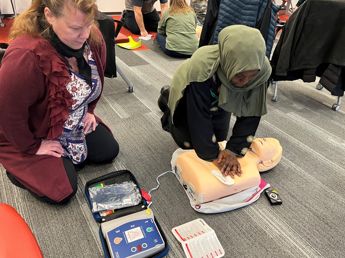 woman performs chest compressions on a manikin while another prepares to set up the AED