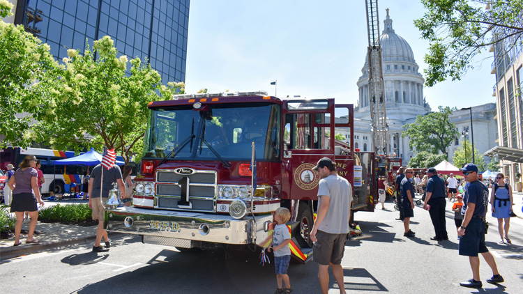 A fire truck with its doors open is parked on the street for the Safety Saturday event. The Capitol building and another fire truck with the ladder extended are in the background. Several residents and firefighters are walking nearby.