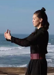 A person standing on a beach with their hands held in front of them. They have light brown skin and shoulder length black hair.