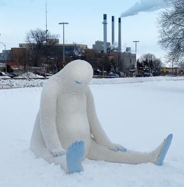 Photograph of the sculpture Sad Yeti (2021) by Actual Size Artworks for winter art carnival Winter is Alive. The white Yeti figure is sitting on the frozen lake with smoke stacks in background.