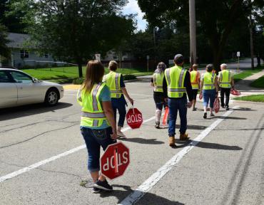 Image shows many crossing guard trainees wearing yellow safety vests, holding hand-held red stop signs, while crossing in a crosswalk.