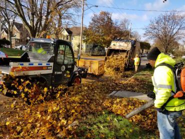 Leaf collection process. Toolcast pushing leaves into collection truck. Laborer using leaf blower. Photo from 2019.