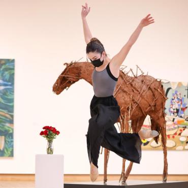 A woman wearing black pants, a grey and black tank top, and a black mask performing a leaping dance pose in front of a gallery of artwork. 