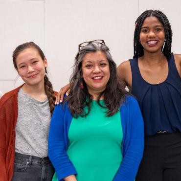 Three figures stand together, Madeleine on the left wears an orange sweater over a grey shirt,  Angie in the middle wears a green dress with a blue sweater, Maliha on the right wears a dark blue tank top