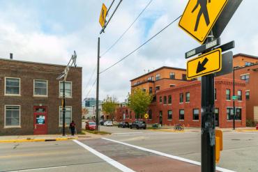 Image shows person with white cane on corner waiting to cross street in crosswalk using an Audible Pedestrian Signal (yellow sign with “walk” figure shown black)