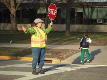 Single crossing guard on duty in crosswalk, with their arms raised, guiding a child across the street while holding a red, hand-held, stop sign. 