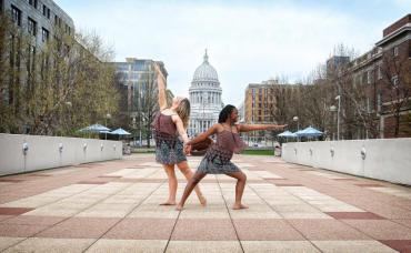 Two dancers from Madison Contemporary Dance pose in the foreground of Monona Terrace with overlapping hands and legs, the state capitol shows in the background