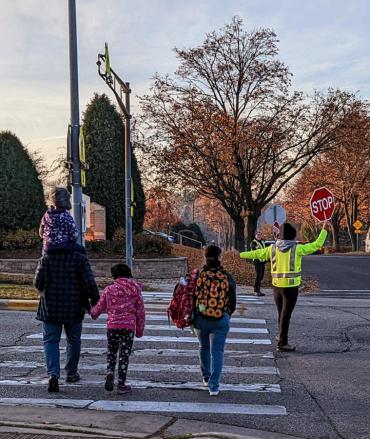 Image of a crossing guard standing in cross walk wearing yellow reflective clothing, holding red stop sign, guiding a family of 3 across street.