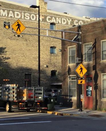 A picture of the accessible pedestrian crossing outside the Wisconsin Council for the Blind.  A brick building with a red door, in from of a 4 story brick building with a ghost sign for Madison Candy Co. on it.