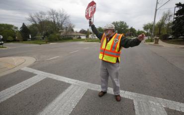 Image of crossing guard in yellow and orange reflective vest holding up red stop sign, standing in crosswalk.