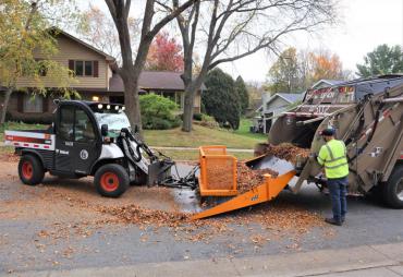 A toolcat is pushing a pile of leaves into a truck. This is the second of the three step process of collecting leaves. First is pulling from the, second is pushing into the truck, and third is street sweeping the leftover debris.