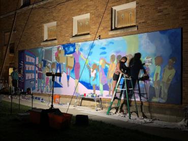 Simone Lawrence Painting Mural on Monroe Street at night with bright lights illuminating wall