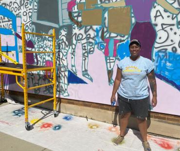 This is a photo of Simone Lawrence standing in front of her in progress mural on Monroe Street