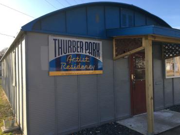 The front of a metal building with a blue semi circular roof, grey walls, and a sign in blue and yellow that reads "Thurber Park Artist Residency." On the right side of the image a rust colored doorway and a small awning are visible.