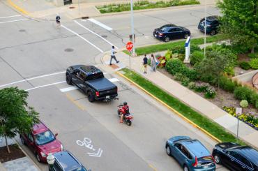 La imagen muestra una intersección de dos caminos, varios vehículos y peatones en el paso de peatones y en las aceras.
