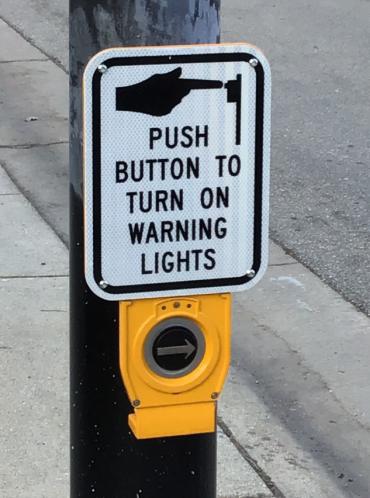 An image of the accessible pedestrian crosswalk button, the sign above it reads, push button to turn on warning lights.