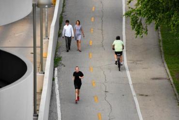 Imagen de un camino de usos múltiples que muestra varios peatones y un corredor en el lado izquierdo del camino dividido que se acerca al espectador, y un ciclista a la derecha que se aleja del espectador.