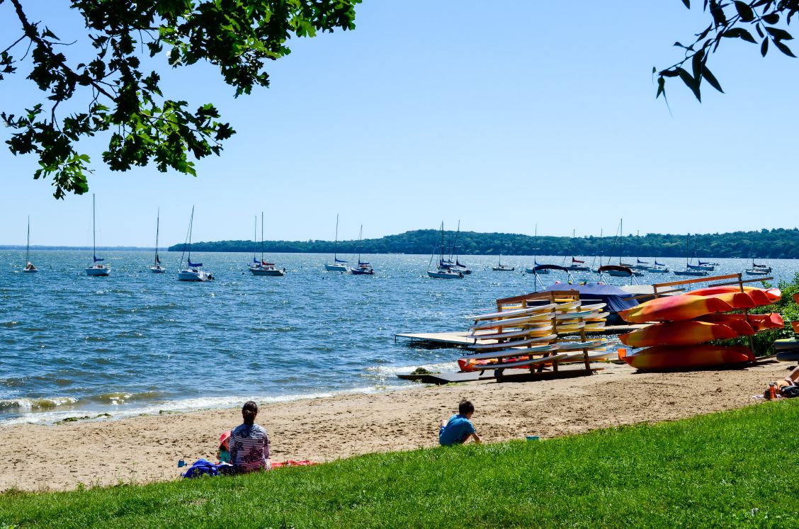 An adult and two children sit on the beach overlooking the lake with sailboats in the distance.